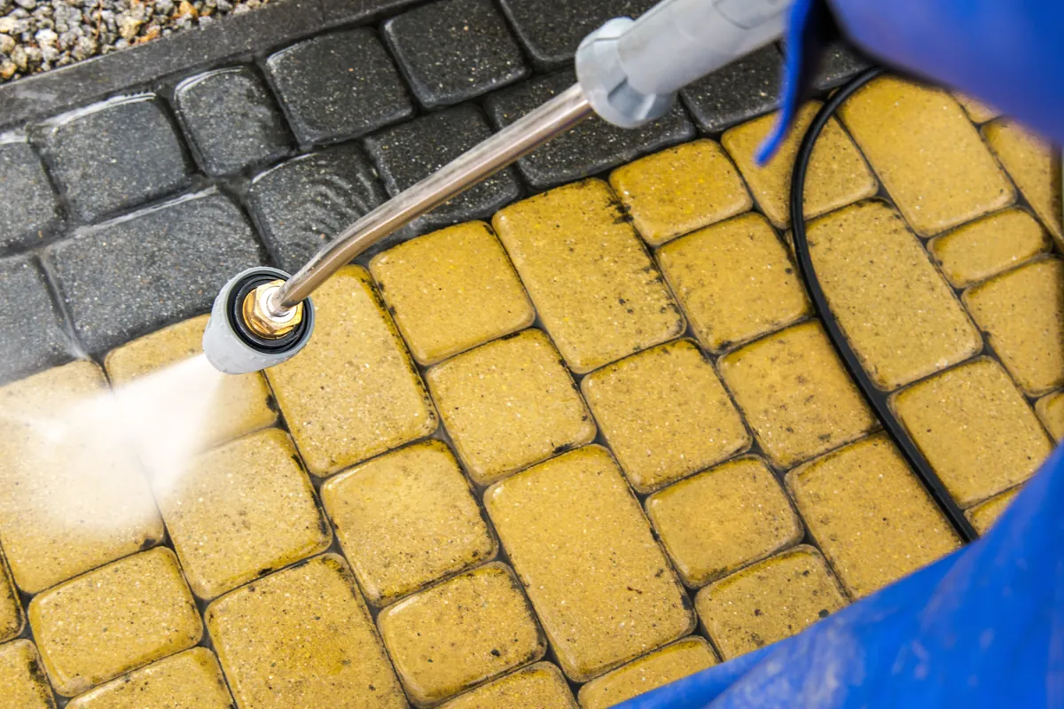 Pressure washer lifting dirt off paver stones, the cleaned strip clearly lighter than the rest.