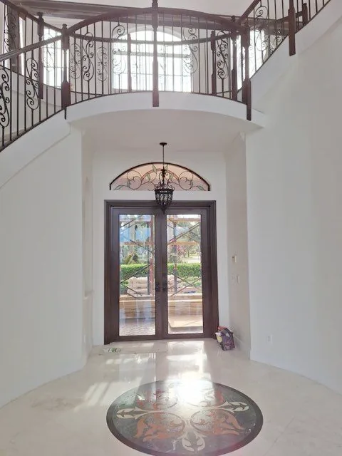 Double-height entry hall painted in a soft white, with an iron-railed landing above and a tiled floor medallion below.